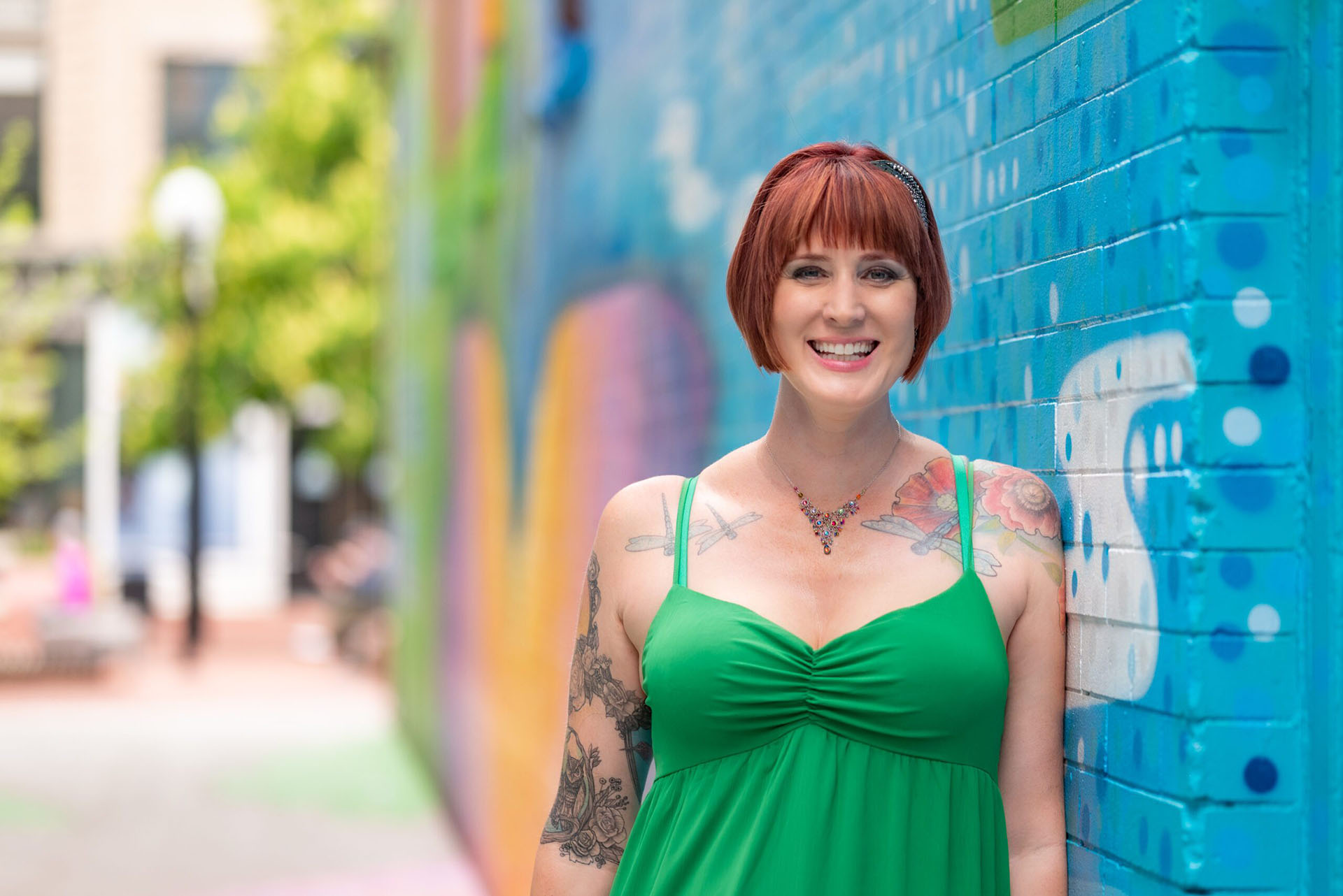 Stephanie Sprenger is wearing a green dress that lets her dragonfly tattoos show, standing in front of a mural.